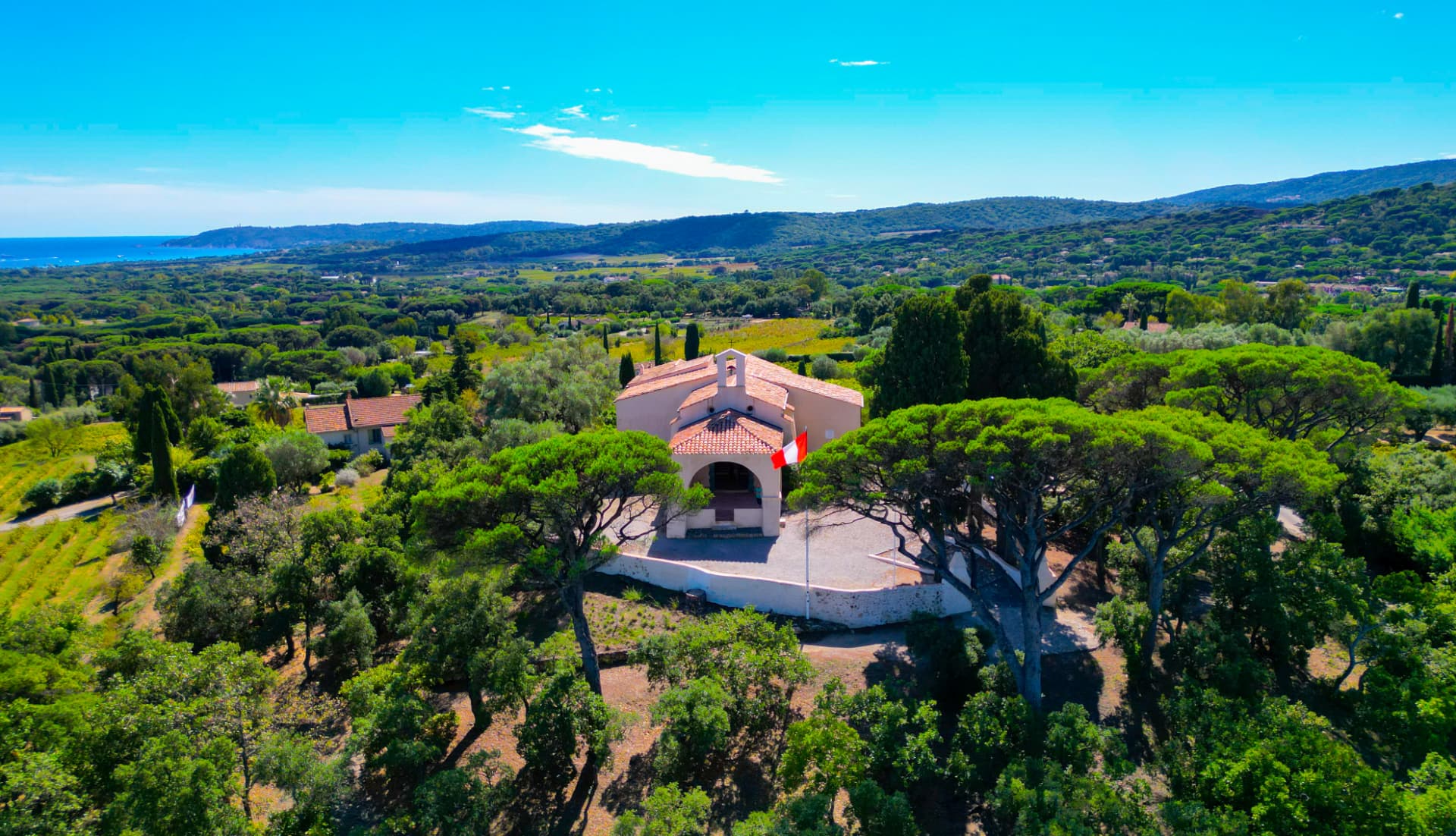 Vue aérienne d'une chapelle entourée de pins et de collines descendant vers la mer